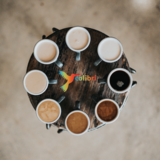 Ein runder, brauner Tisch. Darauf im Kreis Kaffeetassen, gefüllt mit unterschiedlich starkem Kaffee. In der Mitte ein bunter Colibri-Vogel mit dem Schriftzug "Colibri".