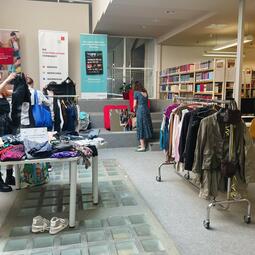 Vergrößerte Ansicht von Foto einer Kleidertauschparty in der Zentralbibliothek. Man sieht Tische und Kleiderstangen voller Kleidung.