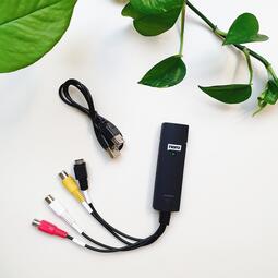 Vergrößerte Ansicht von Zu sehen ist der schwarze Recode Stick mit einem zusätzlichen Kabel. Mit grünen Pflanzenblättern im Hintergrund.