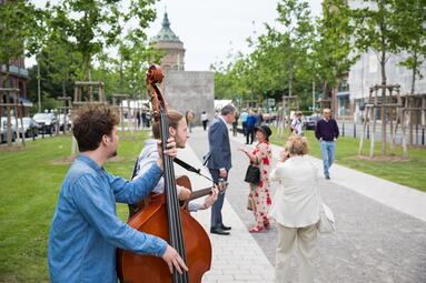Vergrößerte Ansicht von Einweihung der neuen Augusta-Anlage. Im Vordergrund ein Kontrabassist und ein Gitarrist, im Hintergrund Gäste der Feier, die jungen Platanen und der Wasserturm. 