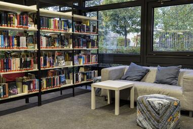 Vergrößerte Ansicht von Ein Foto aus der Zweigstelle Vogelstang. Neben einer Regalwand mit Büchern steht ein graues Sofa, davor ein weißer Beistelltisch und ein quadratischer Hocker.