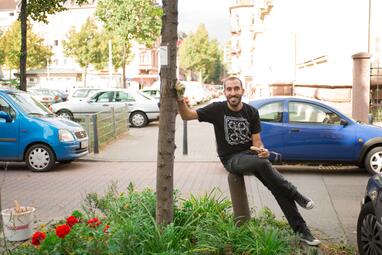 Vergrößerte Ansicht von Ein Mann lehnt an einem Baumstamm, in der Hand ein Gartenwerkzeug. Die Baumscheibe ist üppig bewachsen.