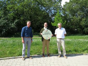Vergrößerte Ansicht von Vor dem Grünstreifen an der Herzogenriedstraße stehen Markus Roeingh, Leiter des Stadtraumservice Mannheim, daneben Bürgermeister Dirk Grunert mit dem Schild "Goldene Wildbiene", daneben Klaus Schwennen, Abteilungsleiter Grünflächen beim Stadtraumservice.