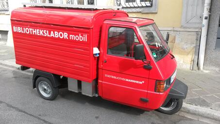 Vergrößerte Ansicht von Foto des mobilen Bibliothekslabors, einer roten Ape, parkend am Straßenrand