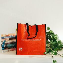 Vergrößerte Ansicht von Foto einer roten Tragetasche, daneben ein Stapel Bücher, rechts eine Pflanze