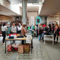 Vergrößerte Ansicht von Foto einer Kleidertauschparty in der Zentralbibliothek. Man sieht Tische voller Kleidung und stöbernde Menschen.