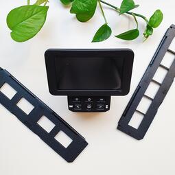 Vergrößerte Ansicht von Man sieht ein schwarzes Gerät, mit dem man Dias scannen kann. Dabei sind zwei Leisten, in die man das zu scannende Material einlegen kann. Mit grünen Pflanzenblättern im Hintergrund.