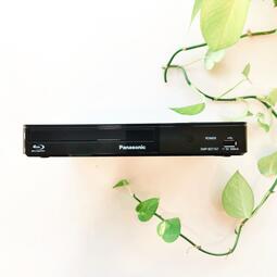 Vergrößerte Ansicht von Frontansicht eines schwarzen Blu-ray-Players. Weißer Hintergrund mit grüner Pflanze.