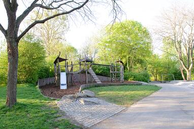 Vergrößerte Ansicht von Ein Spielplatz am Vogelstang-See, umgeben von Bäumen. Man sieht drei Holztürme, die durch Kletter-Elemente miteinander verbunden sind. Ein Turm hat eine Rutsche. Es gibt Netze und Ringe zum Hangeln. 