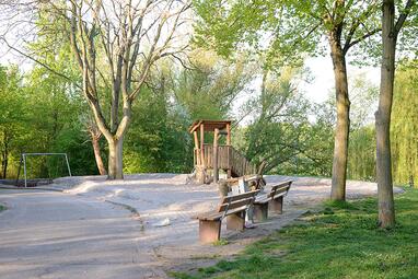Vergrößerte Ansicht von Ein Spielplatz am Vogelstang-See, umgeben von Bäumen. Im Vordergrund zwei Sitzbänke, dahinter ein Kletterturm aus Holz. Im Hintergrund eine Schaukel.