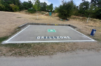 Vergrößerte Ansicht von Die Grillzone am Rheinauer See. Ein rechteckiger gepflasterter Bereich auf einer Wiese. In der Mitte ist ein grünes Grill-Symbol auf den Boden aufgemalt. 