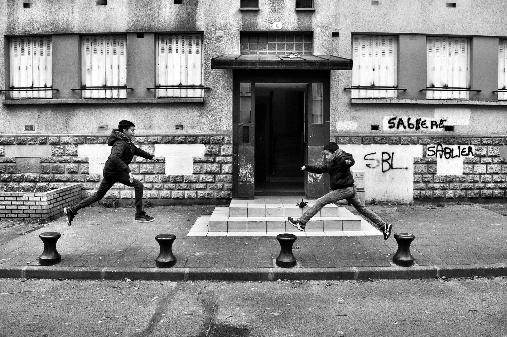 Schwaarz-weiß-Aufnahme zeigt zwei Jungen, die vor einem etwas heruntergekommenen Wohnhauses von Poller zu Poller springen