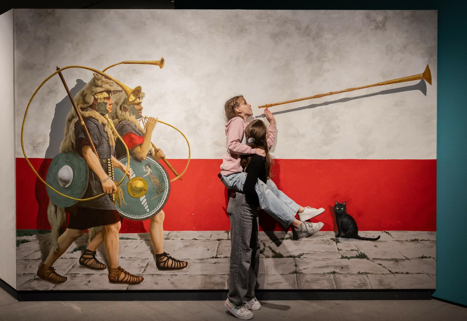 Zwei Kinder posieren vor einem großen Bild, das Legionäre in Uniform beim Marschieren zeigt