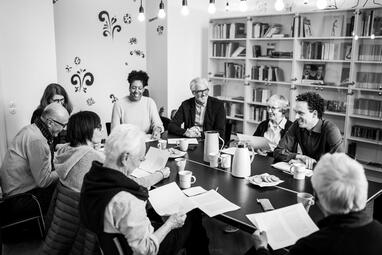 Vergrößerte Ansicht von Foto einer Gruppe Menschen, die mit Texten, Kaffee und Keksen an einem großen Tisch sitzen