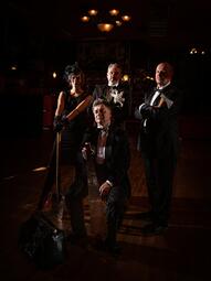 Vergrößerte Ansicht von Dunkles Foto von drei Musikern in schwarzen Anzügen und einer Musikerin in schwarzem Kleid.