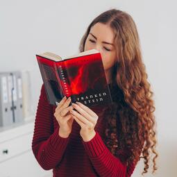 Vergrößerte Ansicht von Foto einer jungen Frau mit langen, gelockten Haaren, die in einem hellen Raum steht und in einem Buch liest. Auf dem Cover steht &quot;Frankenstein&quot;.