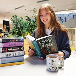 Vergrößerte Ansicht von Foto einer jungen Frau, die in der Bibliothek sitzt und das Buch &quot;Prison Healer&quot; in der Hand hält. Sie lächelt in die Kamera. Neben ihr befinden sich ein Bücherstapel und eine Tasse Tee.