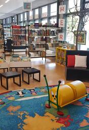 Vergrößerte Ansicht von Foto aus der Zweigstelle Herzogenried, man sieht unter anderem Bücherregale, Sitzgelegenheiten und einen bunten Teppich.