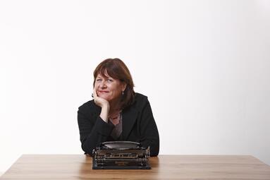 Vergrößerte Ansicht von Foto der Autorin Christine Weiner. Sie sitzt vor einer weißen Wand und an einem braunen Tisch, auf dem eine Schreibmaschine steht.