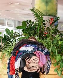 Vergrößerte Ansicht von Foto, auf dem eine junge Frau einen hohen Stapel an Kleidungsstücken hält, sodass er ihr Gesicht fast vollständig verdeckt. Im Hintergrund sieht man grüne Pflanzen.