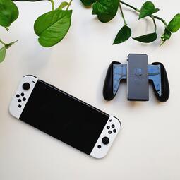 Vergrößerte Ansicht von Zu sehen ist die Nintendo Switch und der dazu gehörende Controller. Mit grünen Pflanzenblättern im Hintergrund.