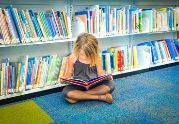 Vergrößerte Ansicht von Kind liest auf dem Boden sitzend vor einem Bücherregal