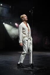 Vergrößerte Ansicht von Eddie Irle steht im weißen Anzug vor einem Standmikrofon, welches zu hoch für ihn ist; er schaut hoch zum Mikrofon.