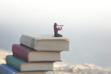 Vergrößerte Ansicht von Auf einem Bücherstapel sitzende Miniaturfigur mit einem Fernglas in der Hand