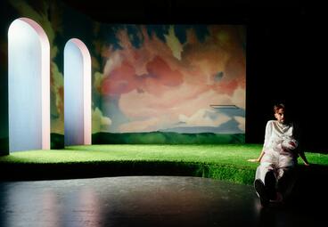 Vergrößerte Ansicht von Auf der Bühne: Kunstrasen, links zwei Torbögen, rosane Wolken auf der Rückwand. Rechts sitzt ein Schauspieler auf der Kante.