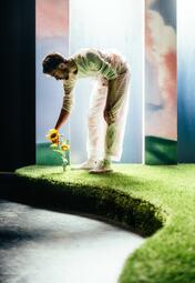 Vergrößerte Ansicht von Ein Schauspieler (Moritz Andrea Bürge) steht vor einem Torbogen und beugt sich hinunter zu einer Sonnenblume