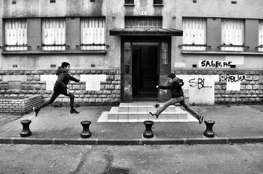 Vergrößerte Ansicht von Schwaarz-weiß-Aufnahme zeigt zwei Jungen, die vor einem etwas heruntergekommenen Wohnhauses von Poller zu Poller springen