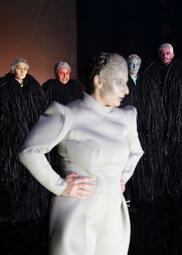 Vergrößerte Ansicht von Im Vordergrund steht eine Person in einem weißen, scharf geschnittenen Kleid. Hinter ihr stehen vier Personen und beobachten sie. Sie tragen schwarze Mäntel, ihre Gesichter sind unifarben angemalt.
