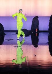 Vergrößerte Ansicht von Larissa Voulgarelis geht mit roboter-tänzerischen Bewegungen auf die Bühne, während im Hintergrund die schwarzen Gestalten ihr Unwesen treiben. Sie trägt einen neongrünen Zweiteiler.