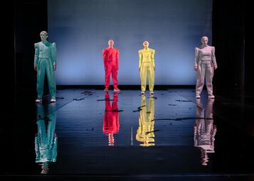 Vergrößerte Ansicht von vier Personen in futuristischen unifarbenen Zweiteilern in unterschiedlichen Farben stehen auf der Bühne