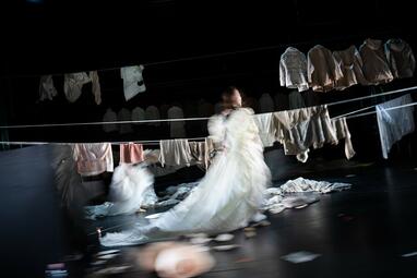 Vergrößerte Ansicht von Mehrere Wäscheleinen mit Kleidungsstücken daran ziehen sich über die Bühne. Eine Person im bauschigen weißen Kleid läuft über die Bühne.