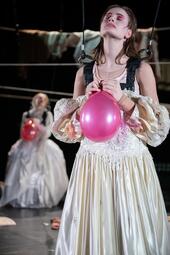 Vergrößerte Ansicht von Antoinette Ullrich trägt ein weißes Kleid und einen schwarzen Brustharness, welcher an zwei von der Decke hängenden Seilen befestigt ist. Sie legt ihren Kopf in den Nacken und schließt die Augen, während sie einen pinken Luftballoon hält.