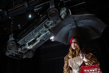 Vergrößerte Ansicht von schräge Froschperspektive. Rahel Weiss trägt einen opulenten Pelzmantel und hält einen schwarzen Regenschirm über ihren Kopf. Über ihr hängt ein schwarzer Mercedes von der Decke.