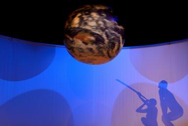 Vergrößerte Ansicht von Carmen Yasemin Zehentmeier und Uwe Topmann schauen sich mit einem Fernrohr die Erde an. Die erde hängt als Kugel mittig von der Decke. Es sind lediglich die Silhouetten der Schauspielenden zu erkennen.