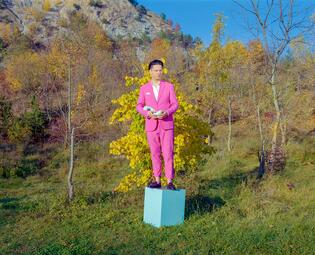 Vergrößerte Ansicht von Konstantin Gropper steht auf einem hellblauen Würfel vor einer herbstlichen Baum- und Berglandschaft. Er trägt einen rosafarbenen Anzug und schaut erst, wärend er ein abstraktes, längliches Objekt hält.