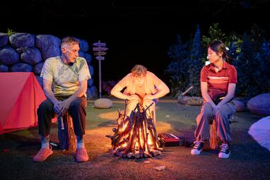 Vergrößerte Ansicht von Uwe Topmann, Rebecca Mauch und Soyi Cho sitzen um ein warm leuchtendes Lagerfeuer und schauen dieses gelangweilt an. Die restliche Bühne ist dunkel; sie ist mit einem orangenen Zelt, einem wegweiser und einigen Felsen ganz im Outdoor-Ferienlager-…
