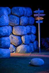 Vergrößerte Ansicht von Eine Felsmauer und ein hölzerner Wegweiser. Hinter der Mauer dringt ein warmes Licht hervor.
