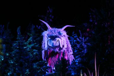 Vergrößerte Ansicht von Rebecca mauch trägt eine schmuckvolle Schafsmaske mit Hörnern. Sie steht zwischen den Nadelbäumen, welche schwach in einem blau-lilafarbenen Licht angeleuchtet werden.