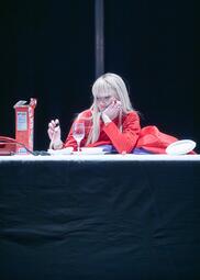Vergrößerte Ansicht von Annemarie Brüntjen trägt eine blonde Perücke und ein rotes Kleid. Sie sitzt an einem weiß gedeckten Tisch mit einer Cornflakes-Packung, Telefon sowie Teller und Besteck. Sie spielt lustlos mit einem Löffel.