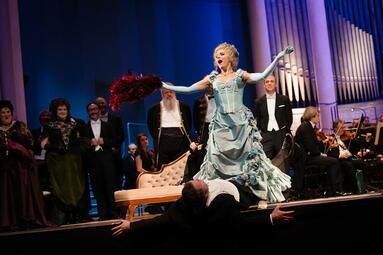 Vergrößerte Ansicht von Amelia Scicolone sitzt auf einer Person, welche fast von der Bühne fällt und erhebt sich mit ausgeweitetnden Armen. Um sie steht der Opernchor und schaut der Szene belustigt zu.