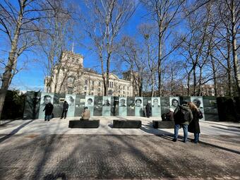 Vergrößerte Ansicht von Das Foto zeigt die im Freien aufgestellten Ausstellungsstücke aus einer gewissen Distanz. Es sind auch interessierte Menschen zu sehen. Im Hintergrund sind Bäume und ein größeres Gebäude.