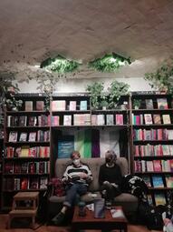 Vergrößerte Ansicht von eine Szene aus einer Lesung. Zwei Personen sitzen auf einem Sofa vor einem Bücherregal. Der Raum ist interessant ausgeleuchtet und Pflanzen stehen zur Dekoration auf den Regalen.