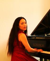 Vergrößerte Ansicht von Ein Foto der Pianistin Sachie Matsushita. Sie sitzt an einem Klavier und hat den Kopf zur Kamera gedreht. Sachie hat glattes schwarzes Haar, das ihr bis zur Hüfte reicht