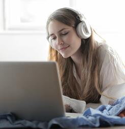 Vergrößerte Ansicht von Eine junge Frau liegt auf dem Bauch und schaut etwas auf ihrem Laptop an. Sie trägt dabei große Kopfhörer.