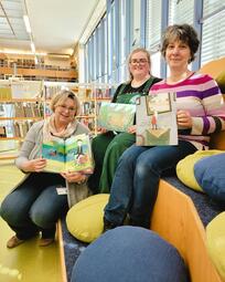Vergrößerte Ansicht von Drei Frauen sitzen auf der Lesetreppe und halten Kinderbücher in die Kamera. Dabei lächeln sie.