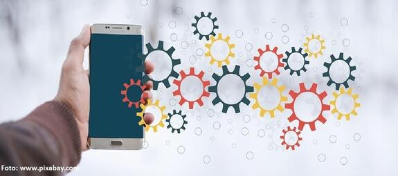 Vergrößerte Ansicht von Eine Hand ragt von links ins Bild und hat ein Smartphone in der Hand. Daneben tauchen bunte Zahnrädchen auf, die die Funktionsweise des Handys symbolisieren sollen.